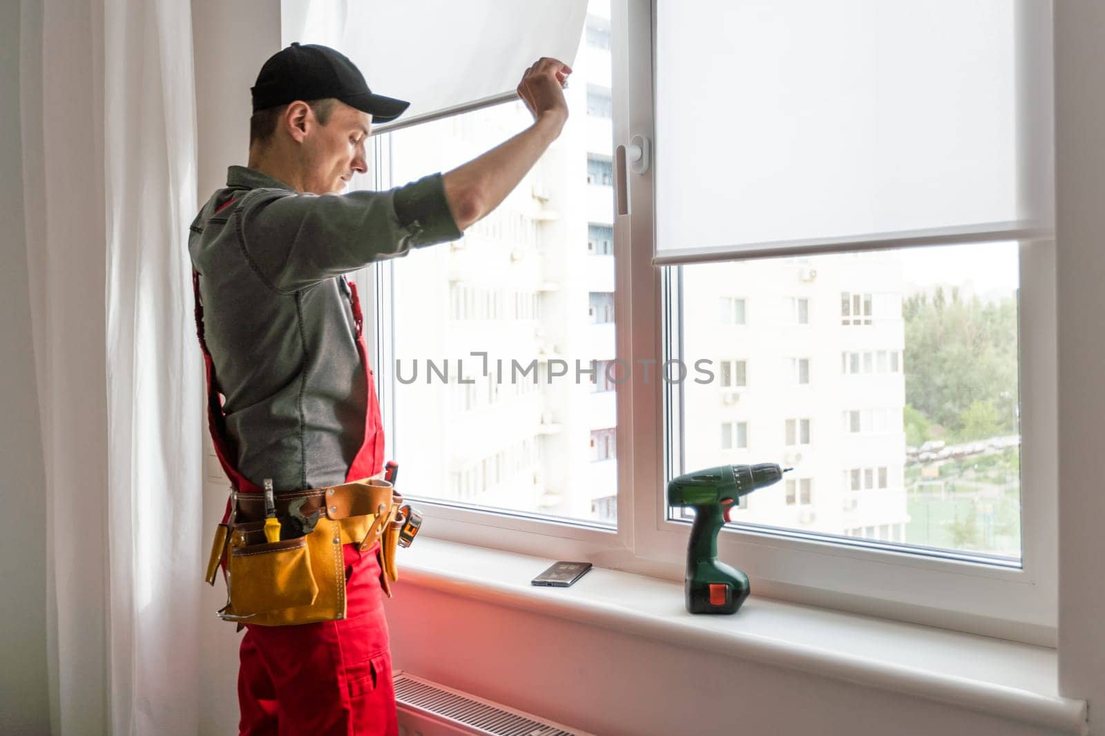 Maintenance man is fixing windows in a living room using a skrewdriver. He is wearing glasses, yellow gloves and a black apron by Andelov13