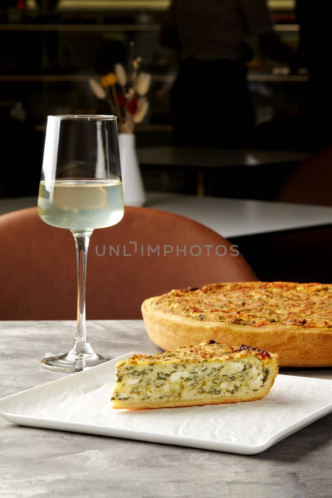 Slice of piquant cheese and spinach quiche with crispy shortbread crust served with glass of white wine in cozy cafe. Elegant dining setting
