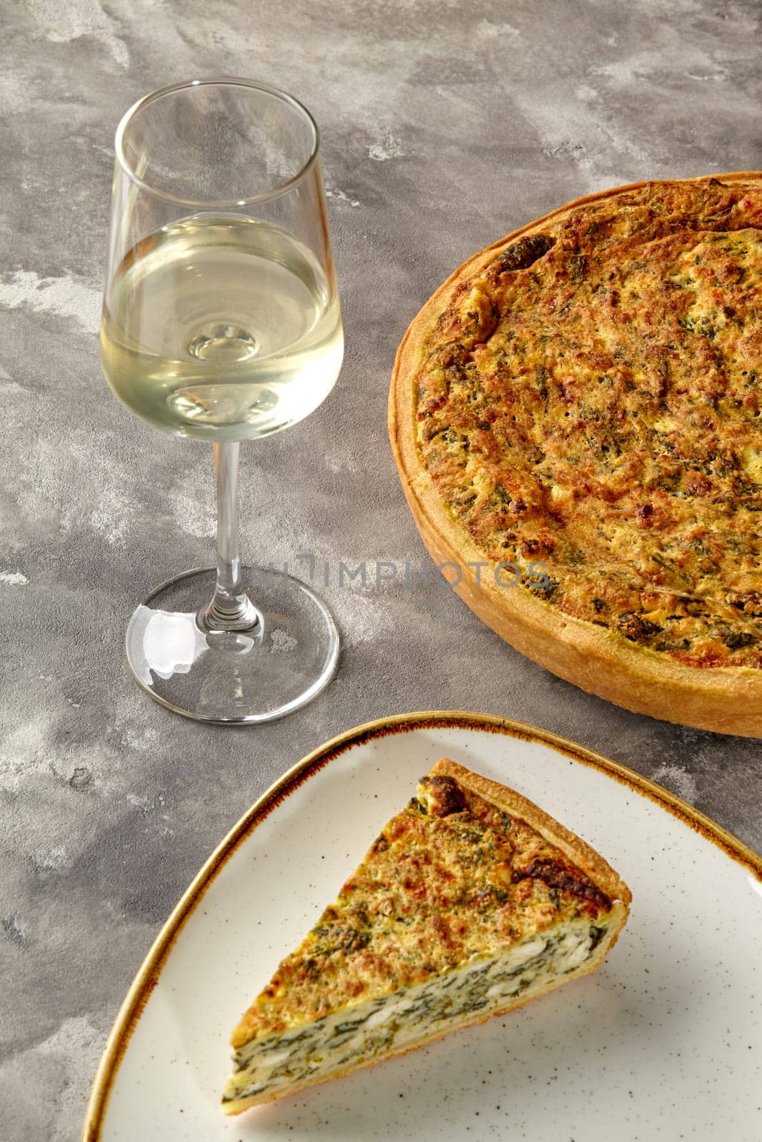 Savoring slice of Quiche Lorraine with cheese and spinach accompanied by glass of chilled white wine, presented on textured background