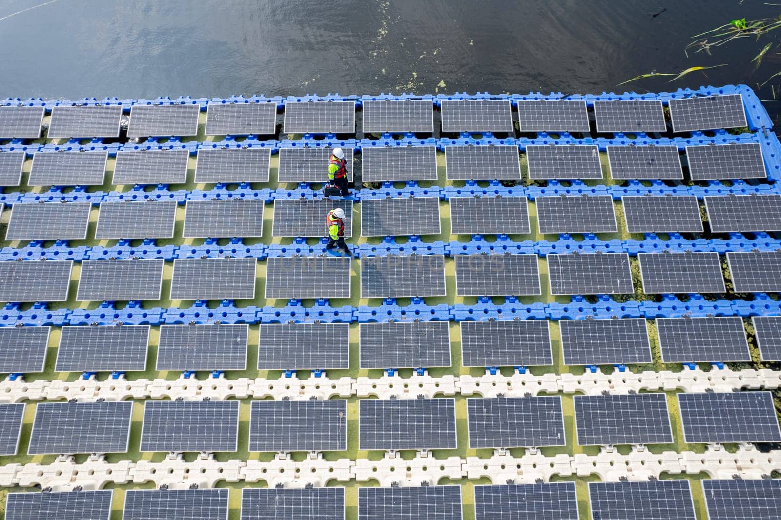 Wide shot of aeroview or top view of two technician workers walk along the solar panel system from left to right side over water reservoir for green ecosystem energy for good environment.