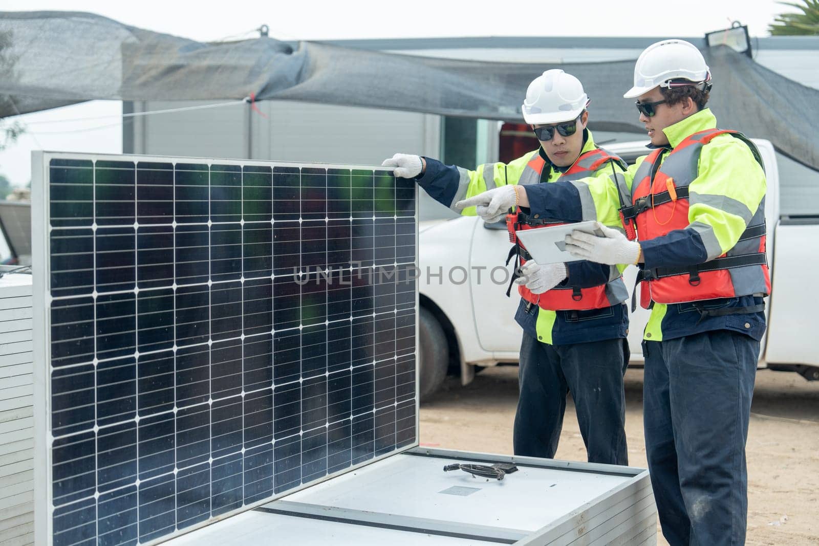 Two Asian professional technician workers hold and discuss using tablet to check solar cell panel and they work in workplace area with concept of green energy system.
