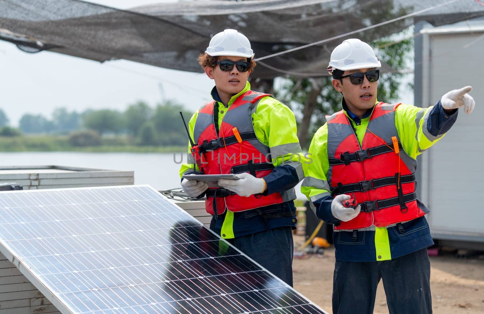 Two technician workers with safety uniform stand beside of solar cell panel and one point forward to discuss about work with his co-worker in workplace. by nrradmin