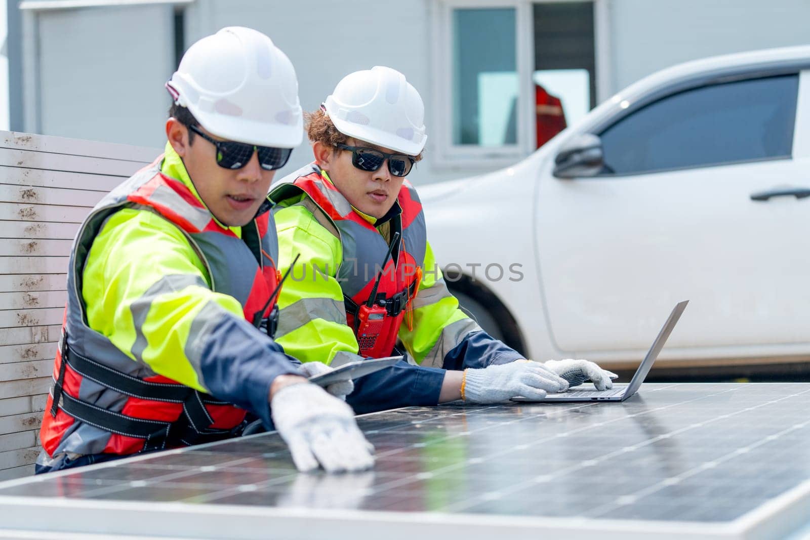 Two Asian professional technician workers sit near solar cell panel and use laptop to discuss about work in concept of green energy system for good environment.