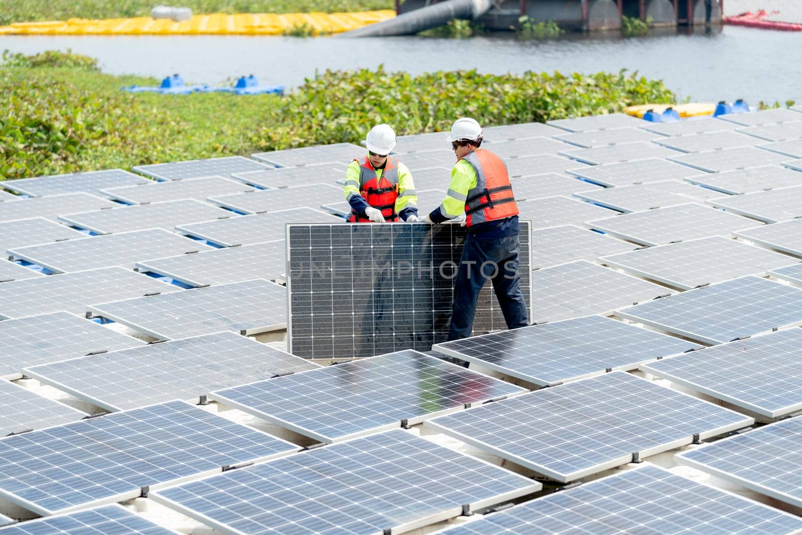 Side view of two Asian professional technician workers with safety uniform stand and help to check or maintenance solar cell panels in area of workplace over water reservoir.