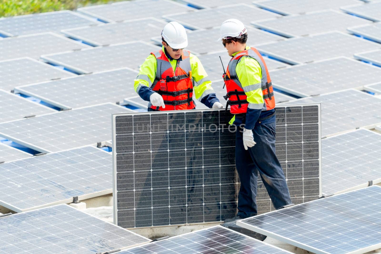 Two Asian professional technician workers with safety uniform stand and help to check or maintenance solar cell panels in area of workplace over water reservoir. by nrradmin
