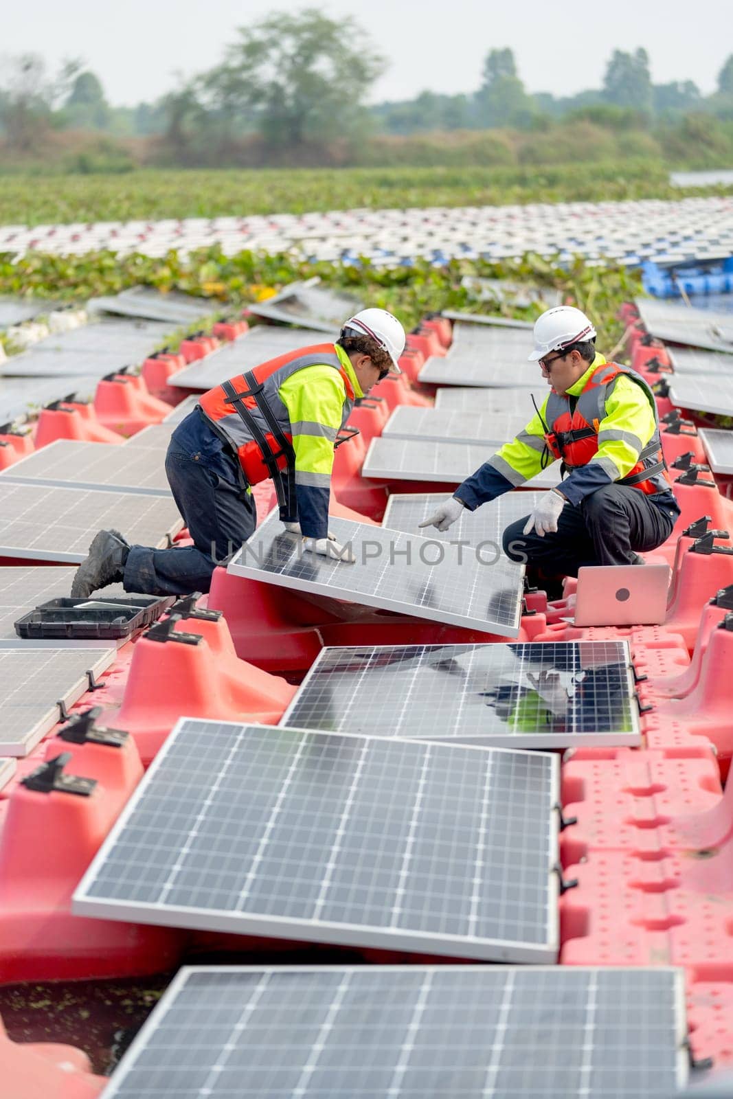 Vertical image of professional technician worker use laptop to work with other co-worker touch to check and maintenance solar cell panel in workplace over water reservoir.