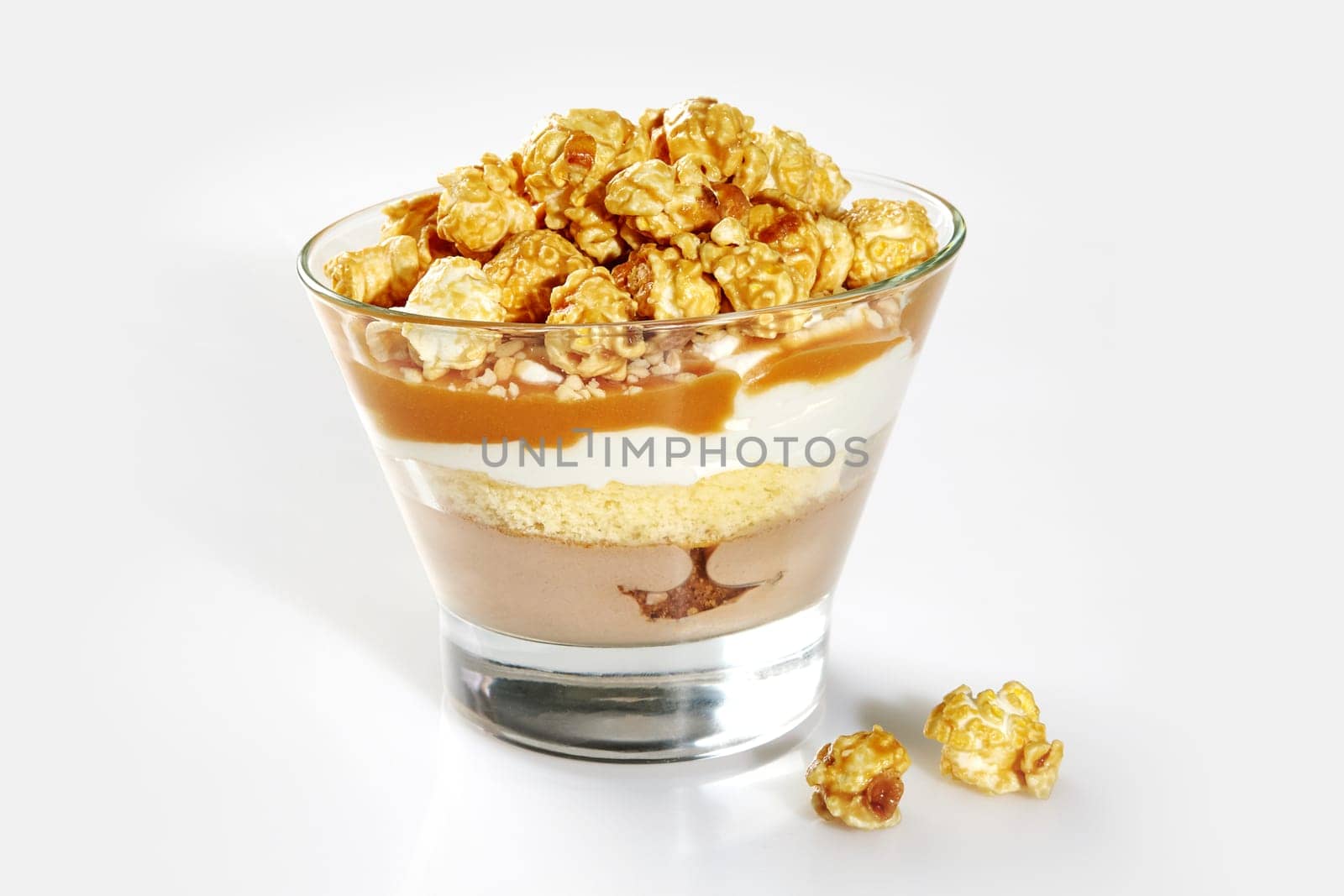 Tempting sweet layered trifle with sponge cake, panna cotta, rich caramel sauce, whipped cream, and caramel coated popcorn in clear dessert glass on white background