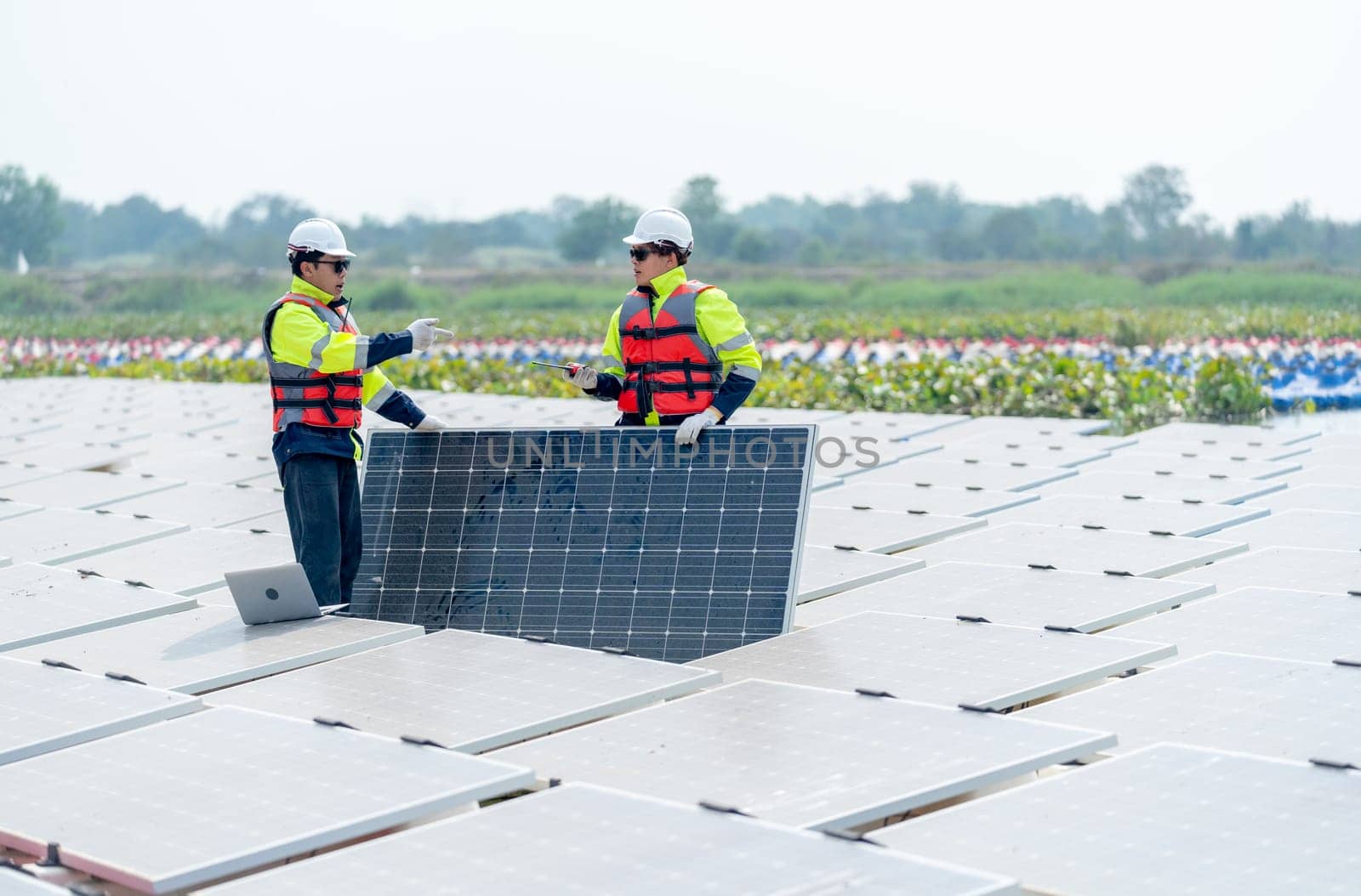 Two professional technician workers stand and hold solar cell panel also discuss together in area of network of base over area of water reservoir.