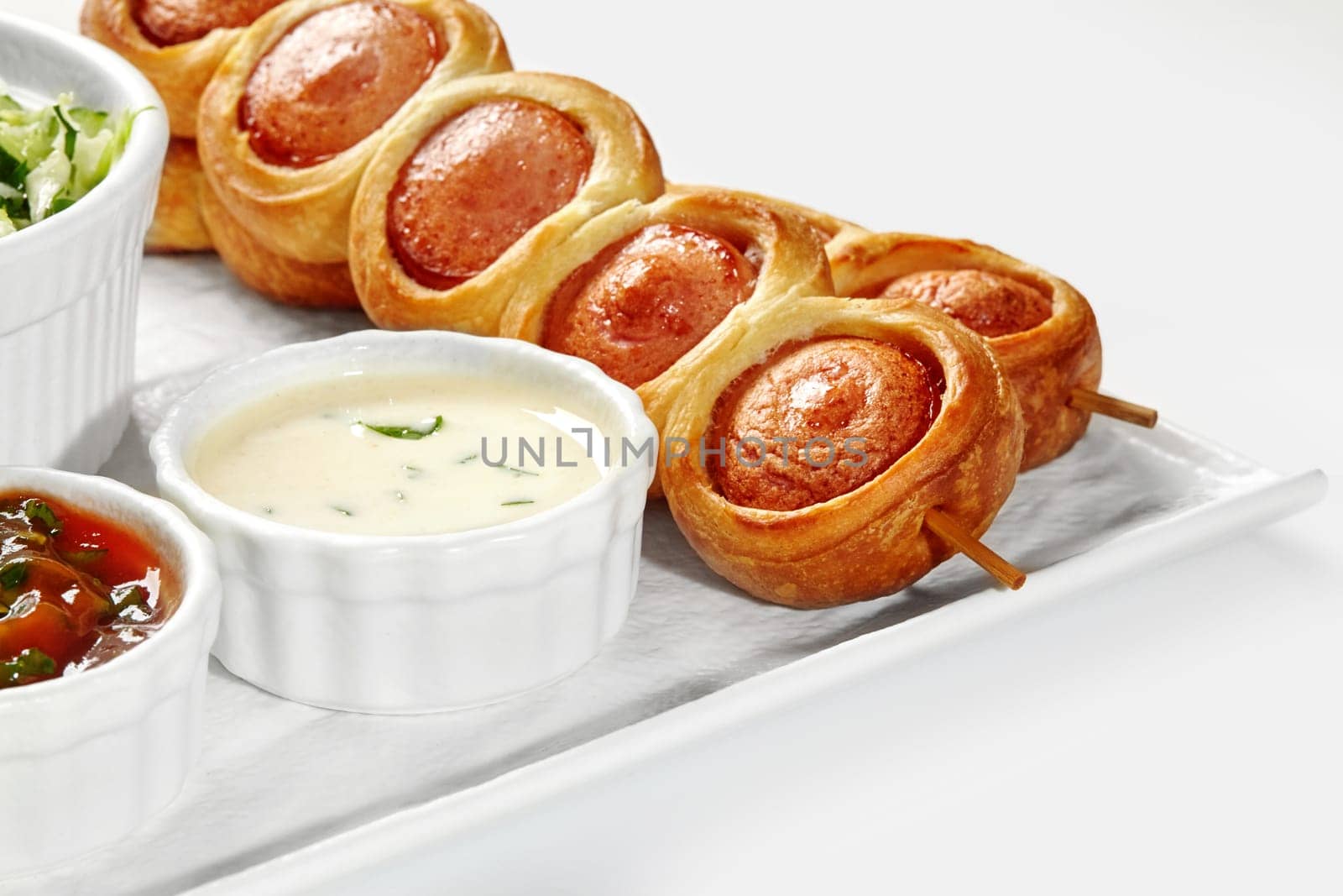 Closeup of appetizing golden brown sausage rolls in brioche on skewers served on plate with vegetable salad and sauces against white background. French style breakfast