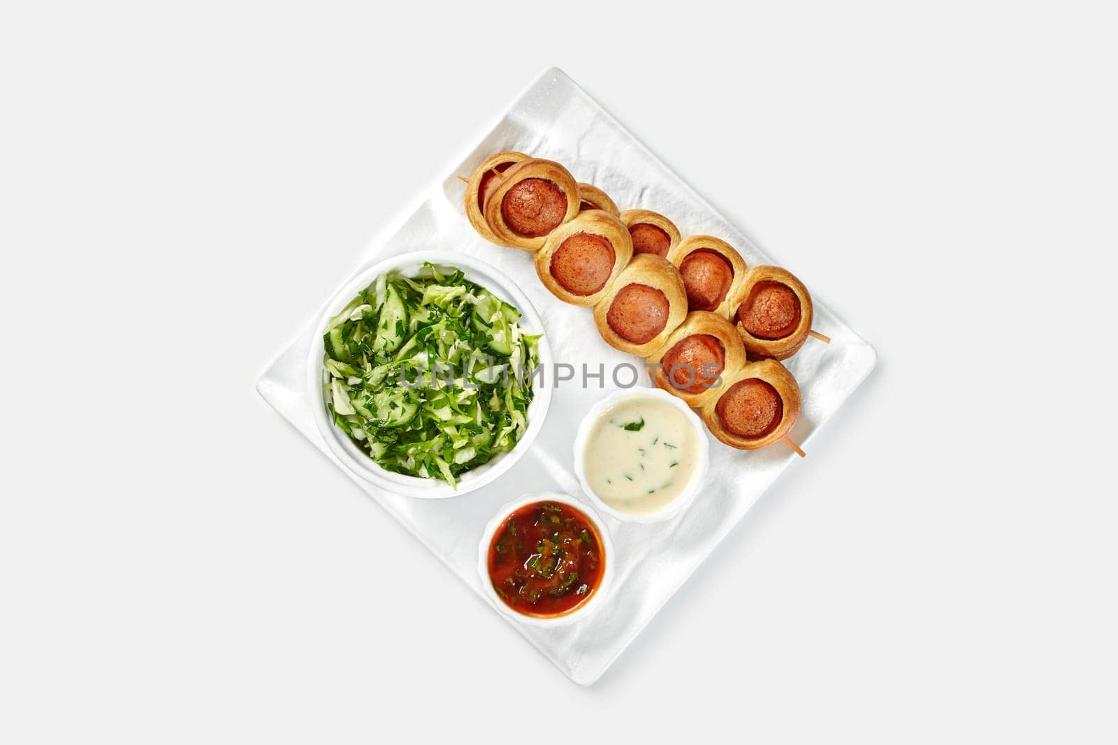 Hearty breakfast set with freshly baked sausage rolls, healthy vegetable crisp salad, and spicy dipping sauces on tray against white backdrop