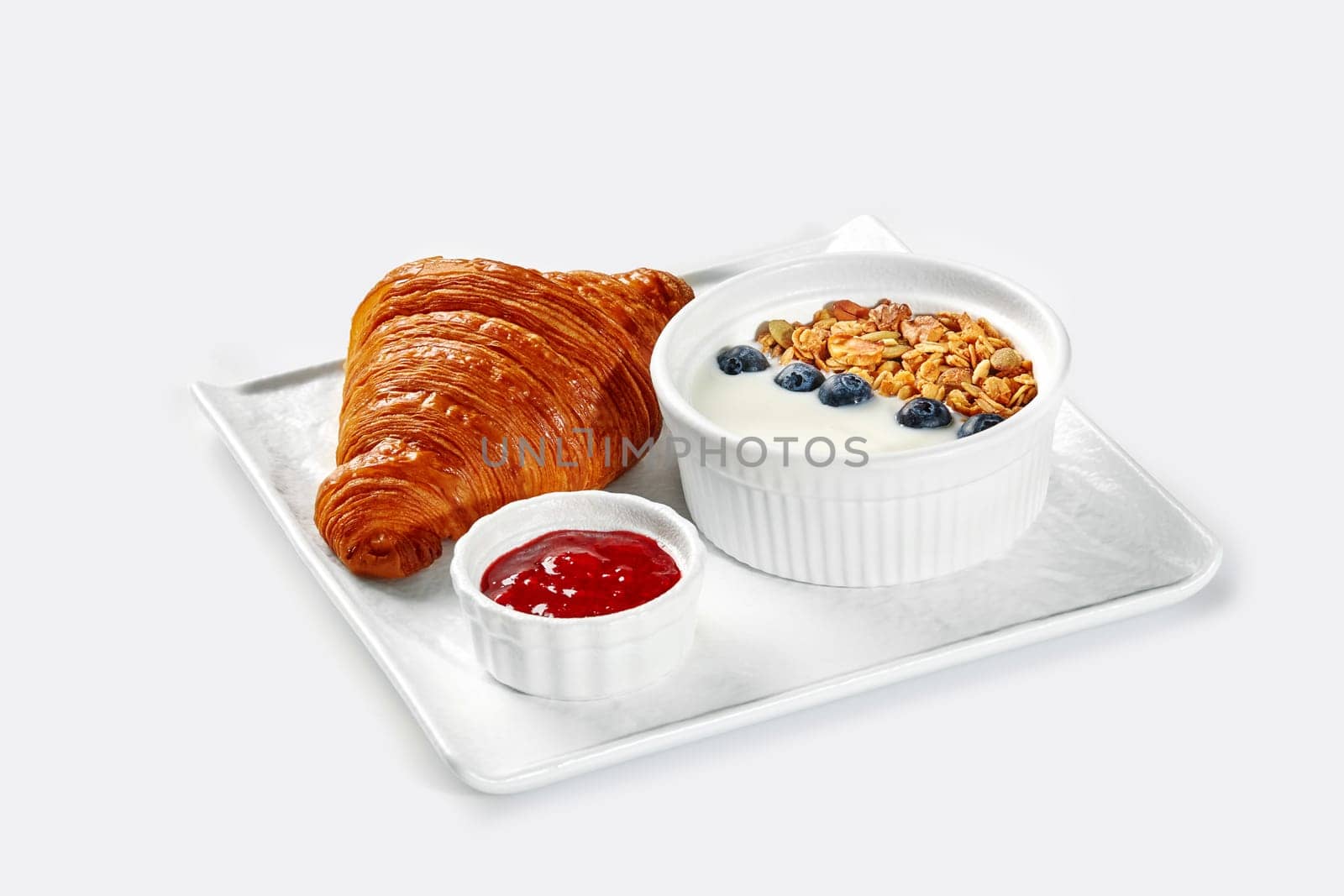 Appetizing breakfast set of golden puff croissant, light creamy yogurt with crispy granola and blueberries, and fresh sweet berry jam on white tray