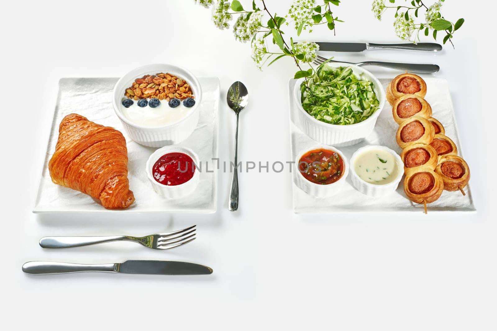 Delightful breakfast arrangement with golden croissant, granola, yogurt, sausages in brioche on skewers, green salad, and dips on clean white setting decorated with tender flowers