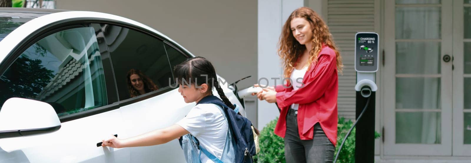 Happy little young girl learn about eco-friendly and energy sustainability as she help her mother recharge electric vehicle from home EV charging station. EV car and modern family. Panorama Synchronos