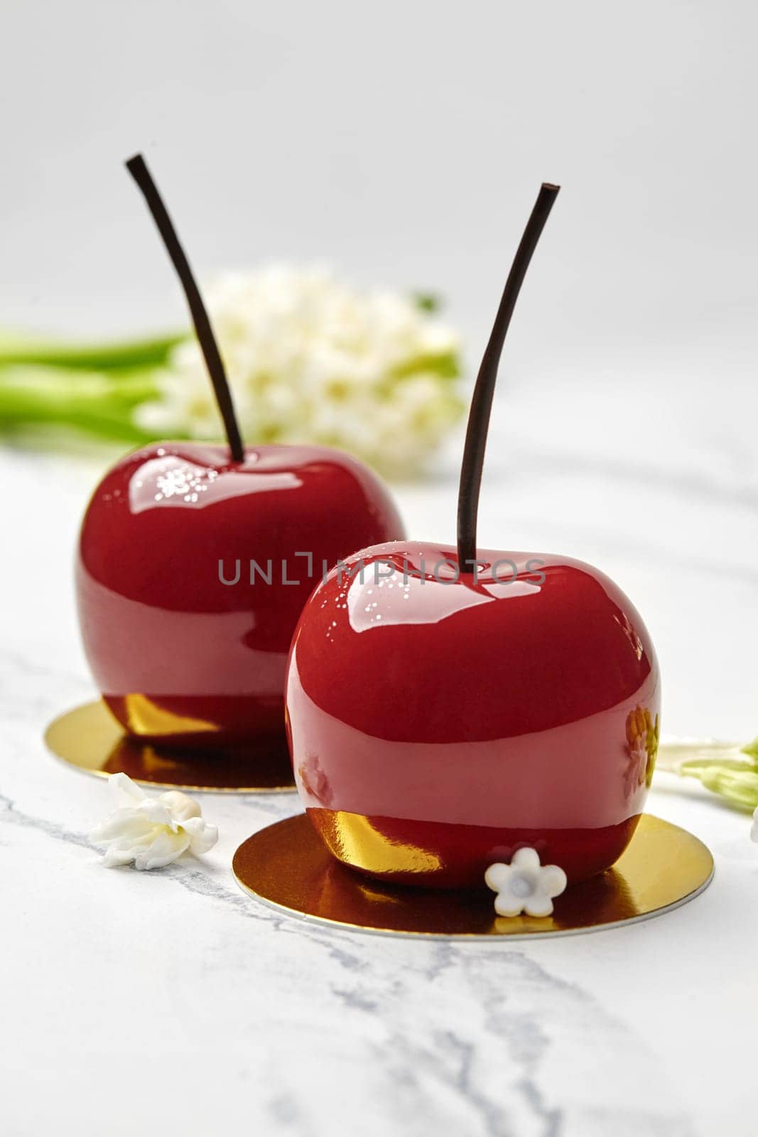 Inspired by beauty of nature, artisan desserts shaped like cherries with glossy red glaze and chocolate stems, presented on marble with gold accents and white flowers in background