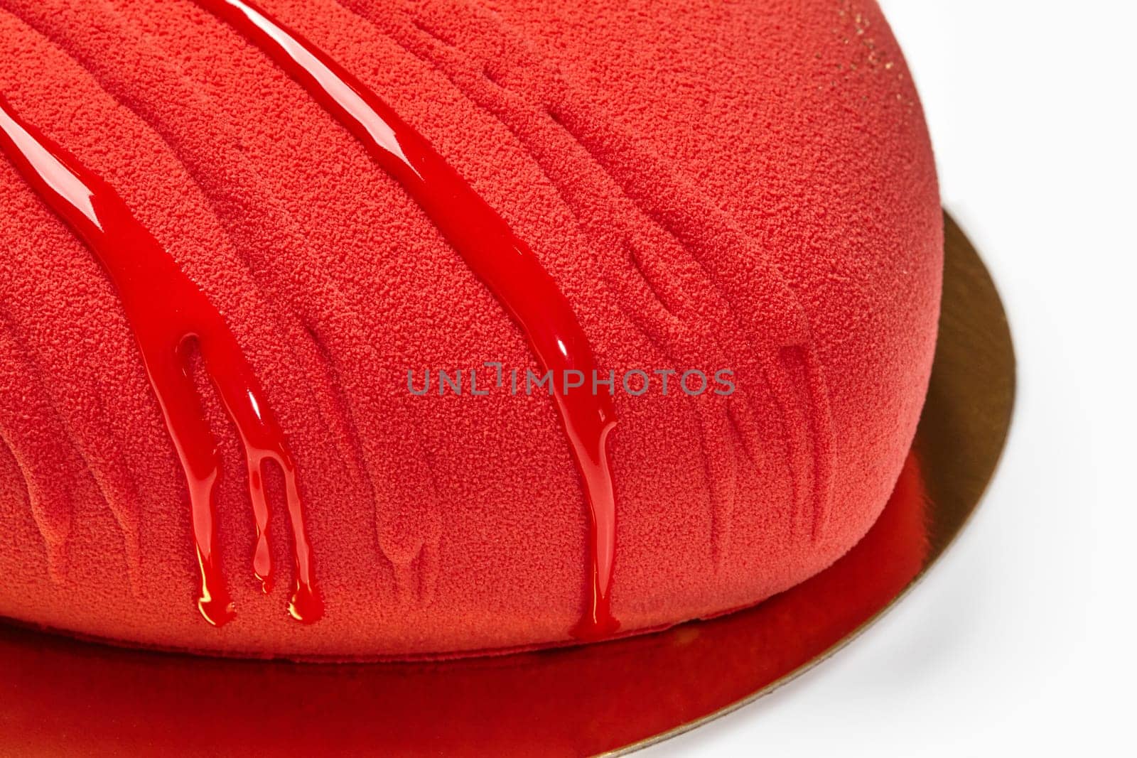 Close-up view of heart-shaped red mousse cake with velvety texture, drizzled with glossy glaze on reflective golden base. Stylish designer dessert