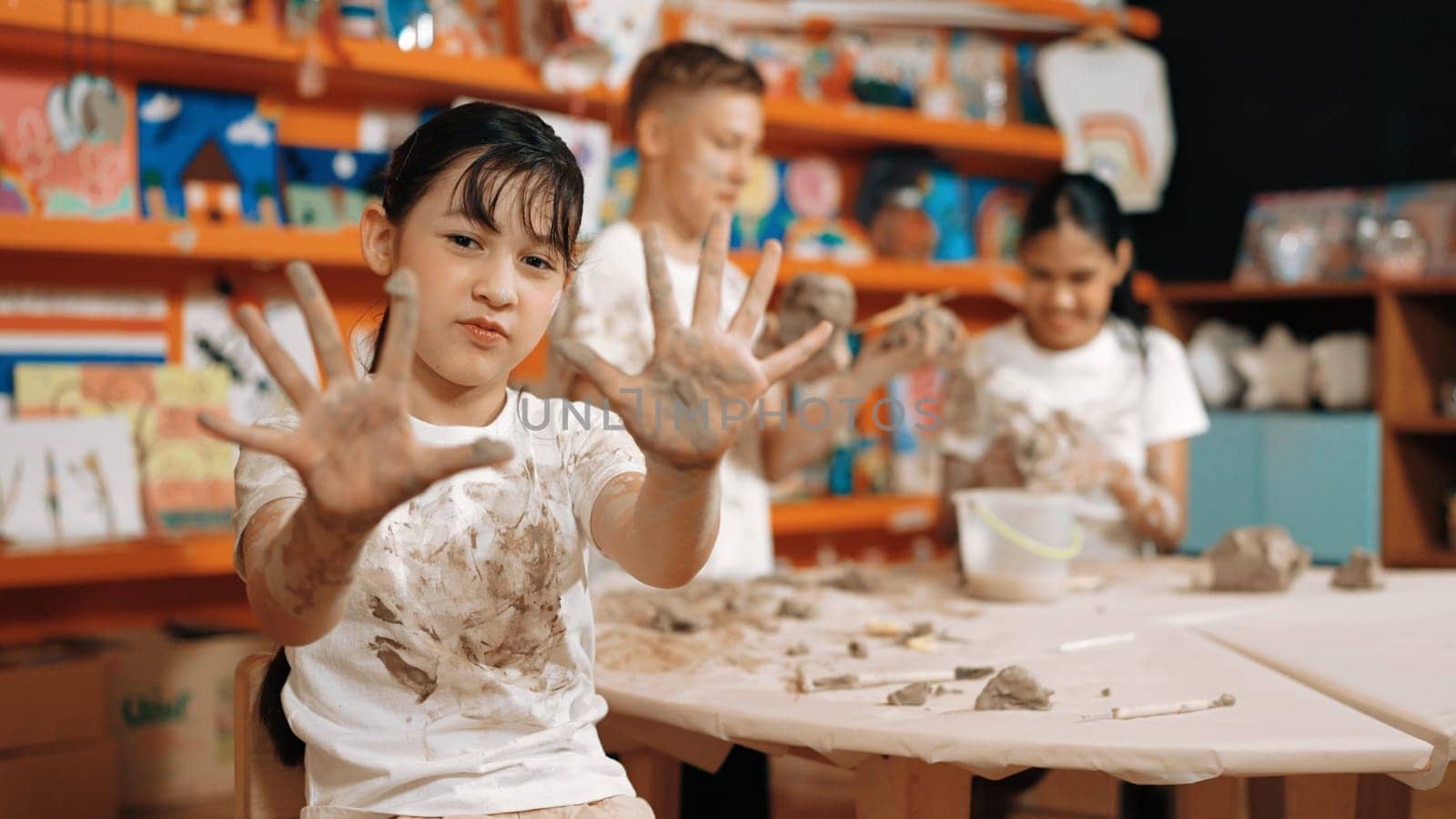 Asian girl showing front hand while highschool children modeling clay. Smart diverse student wearing dirty shirt while looking at camera at workshop in art lesson. Blurring background. Edification.