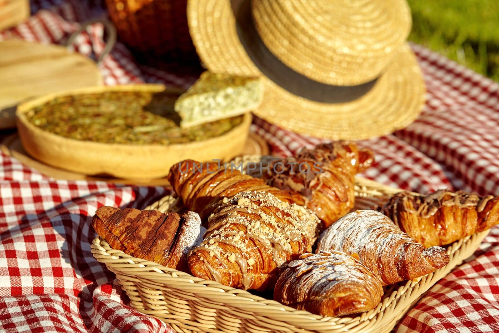 Wicker tray with sweet fluffy puff croissants on red and white checkered blanket on blurred background with savory quiche and straw hat during cozy picnic on sunny summer day