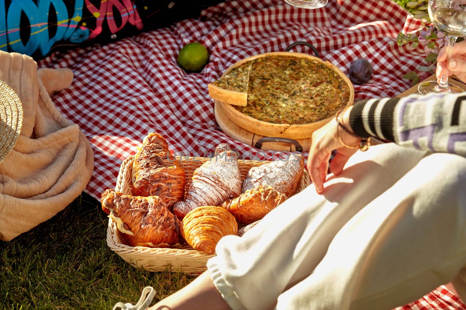 Cozy summer picnic scene on sunny day on green grass with sweet fluffy croissants in wicker basket, homemade savory quiche and fresh fruits scattered on checkered blanket, cropped shot