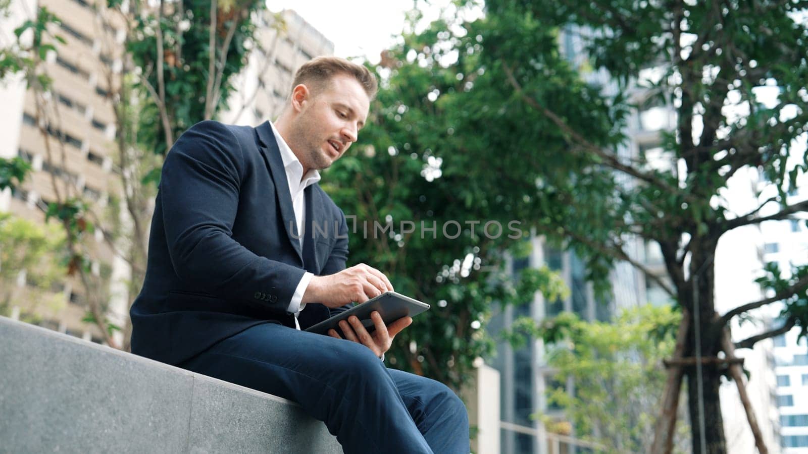 Professional businessman sitting at park while working on phone by reading report from marketing team. Happy manager looking increasing sales or successful project while smiling at eco city. Urbane