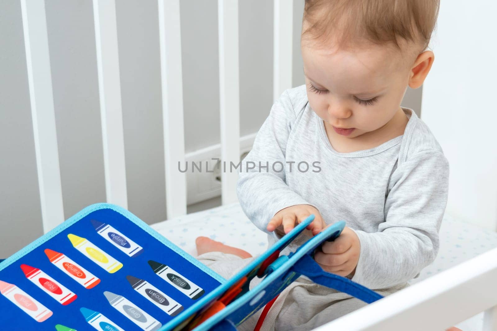 One year old baby playing with montessori busy book sitting in crib