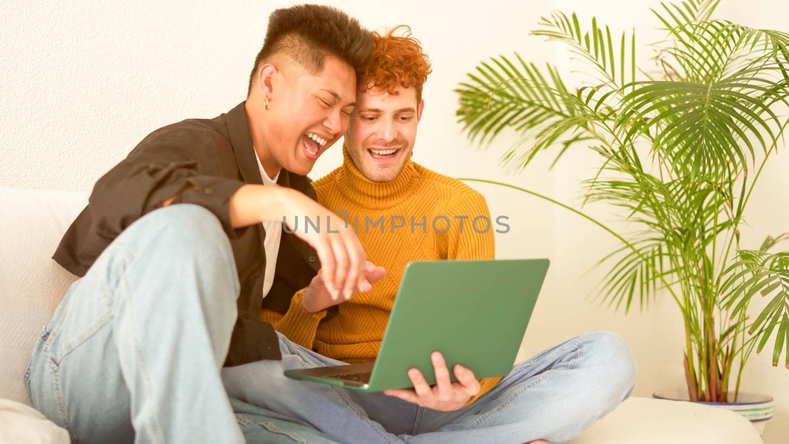 Gay couple laughing while using a laptop sitting on the sofa at home