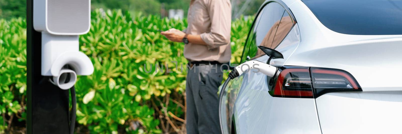 Young man use smartphone to pay for electricity at public EV car charging station green city park. Modern environmental and sustainable urban lifestyle with EV vehicle. Panorama Expedient