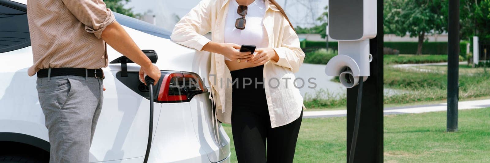 Young couple use smartphone to pay for electricity at public EV car charging station green city park. Modern environmental and sustainable urban lifestyle with EV vehicle. Panorama Expedient