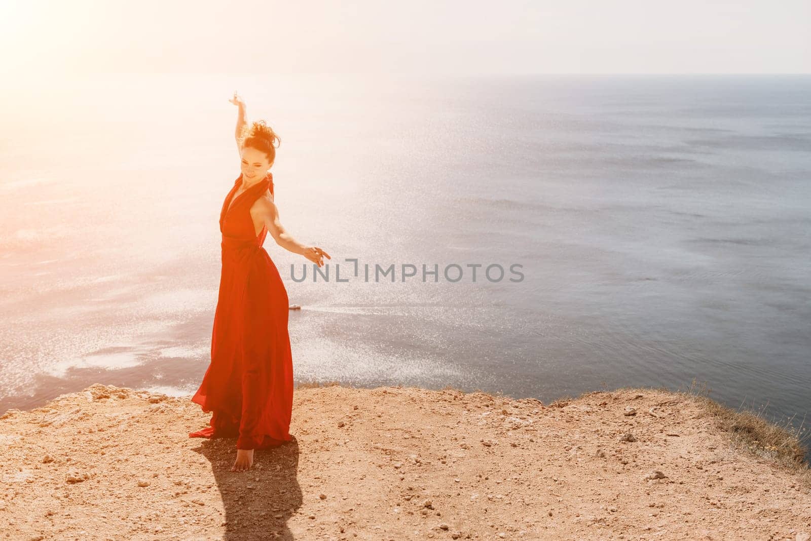 Woman in red dress on sea. Side view a Young beautiful sensual woman in a red long dress posing on a rock high above the sea on sunset. Girl on the nature on blue sky background. Fashion photo. by panophotograph