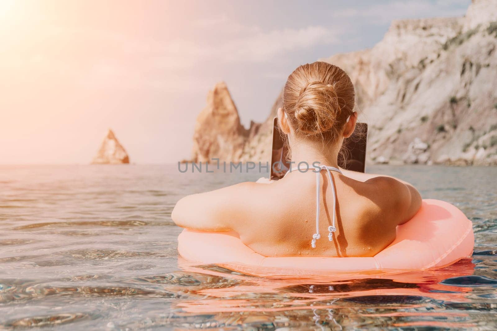 Woman freelancer works on laptop swimming in sea on pink inflatable ring. Pretty lady typing on computer while floating in the sea on inflatable donut at sunset. Freelance, remote work on vacation