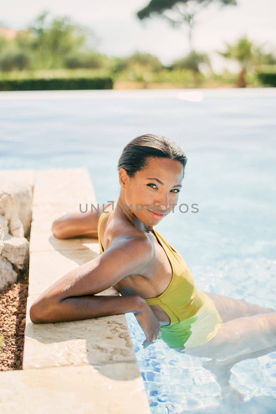 Young smiling woman sits in a swimming pool leaning on the edge and turning her head to the side. High quality photo