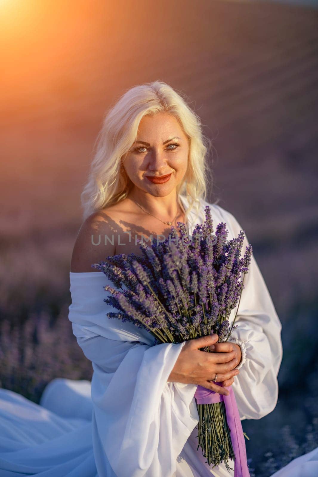 Blonde woman poses in lavender field at sunset. Happy woman in white dress holds lavender bouquet. Aromatherapy concept, lavender oil, photo session in lavender.