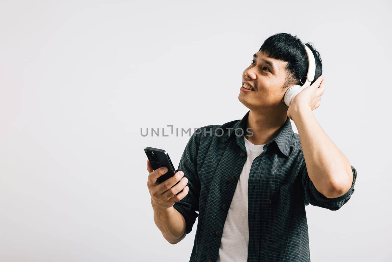 Portrait of a cheerful Asian man singing and listening to music on his smartphone with Bluetooth earphones. Studio shot isolated on white, emphasizing his love for karaoke.