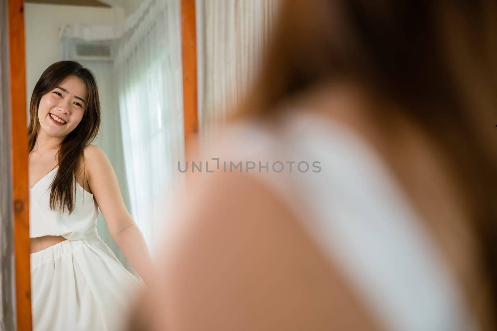 Happy woman wearing dress on front mirror looking clothes at home after morning wake up, Asian beautiful young female trying on blouse and smiling while standing in front of mirror in room