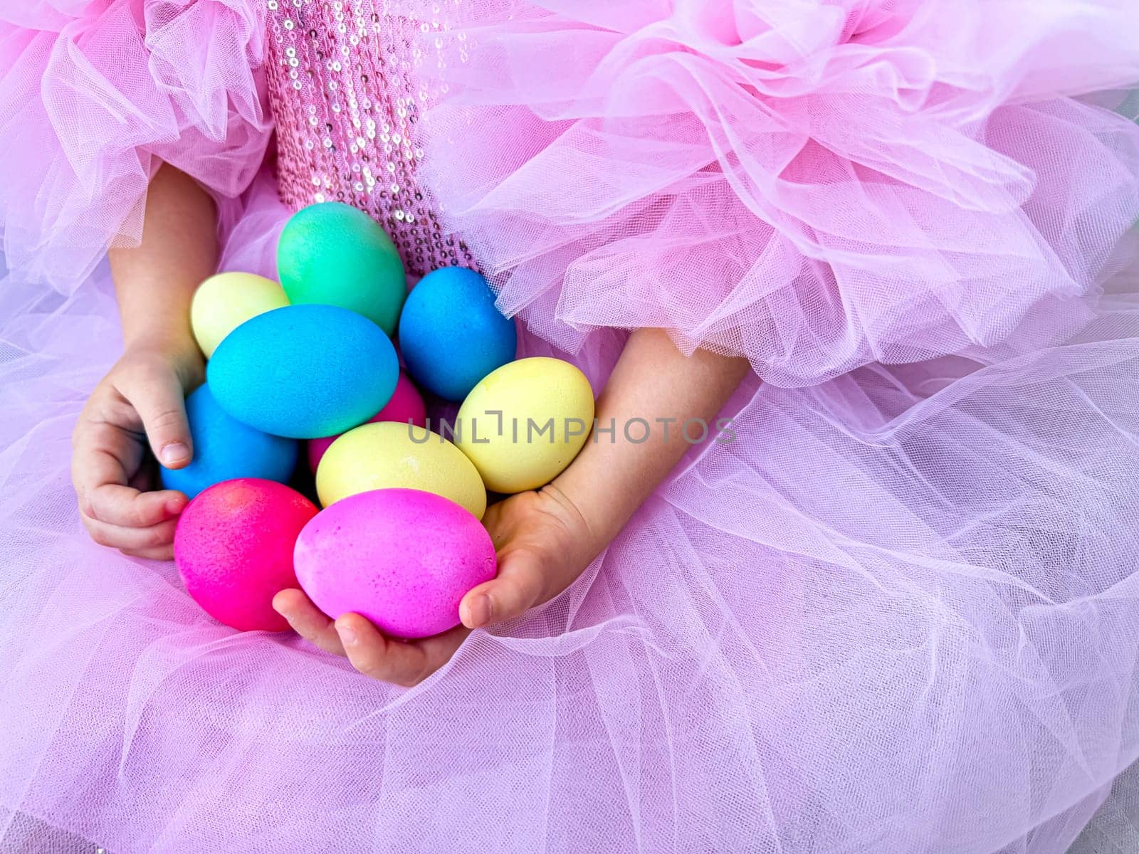 Child in pink tulle dress holds in her hands handful of bright multi colored Easter eggs, symbolizing the spring holidays and the celebration of Easter. by Lunnica