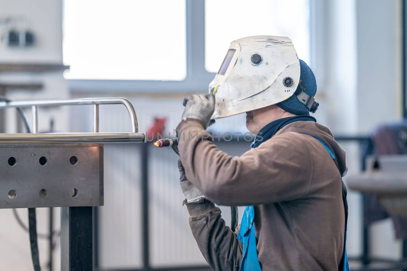 A worker in personal protective equipment, including a helmet and workwear, is welding metal using a gaspowered machine inside a building