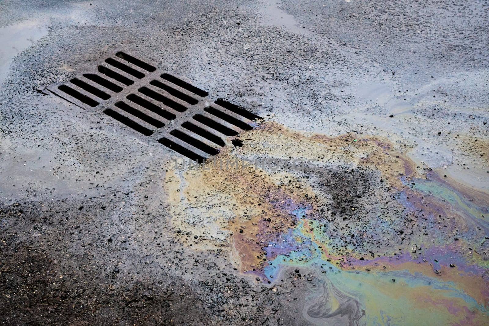 An oil or gasoline slick against the backdrop of an asphalt road flows into a storm drain. Environmental problems of water pollution.