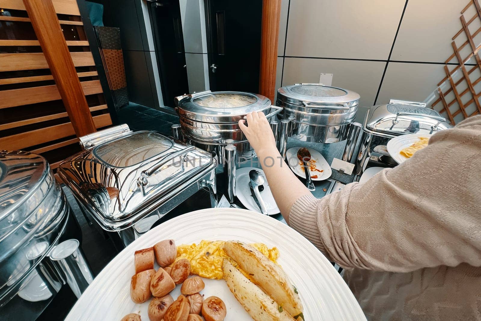 A lady at the hotel buffet is selecting food from a warming machine to place on her plate. A person arranging food on a plate from their own perspective.