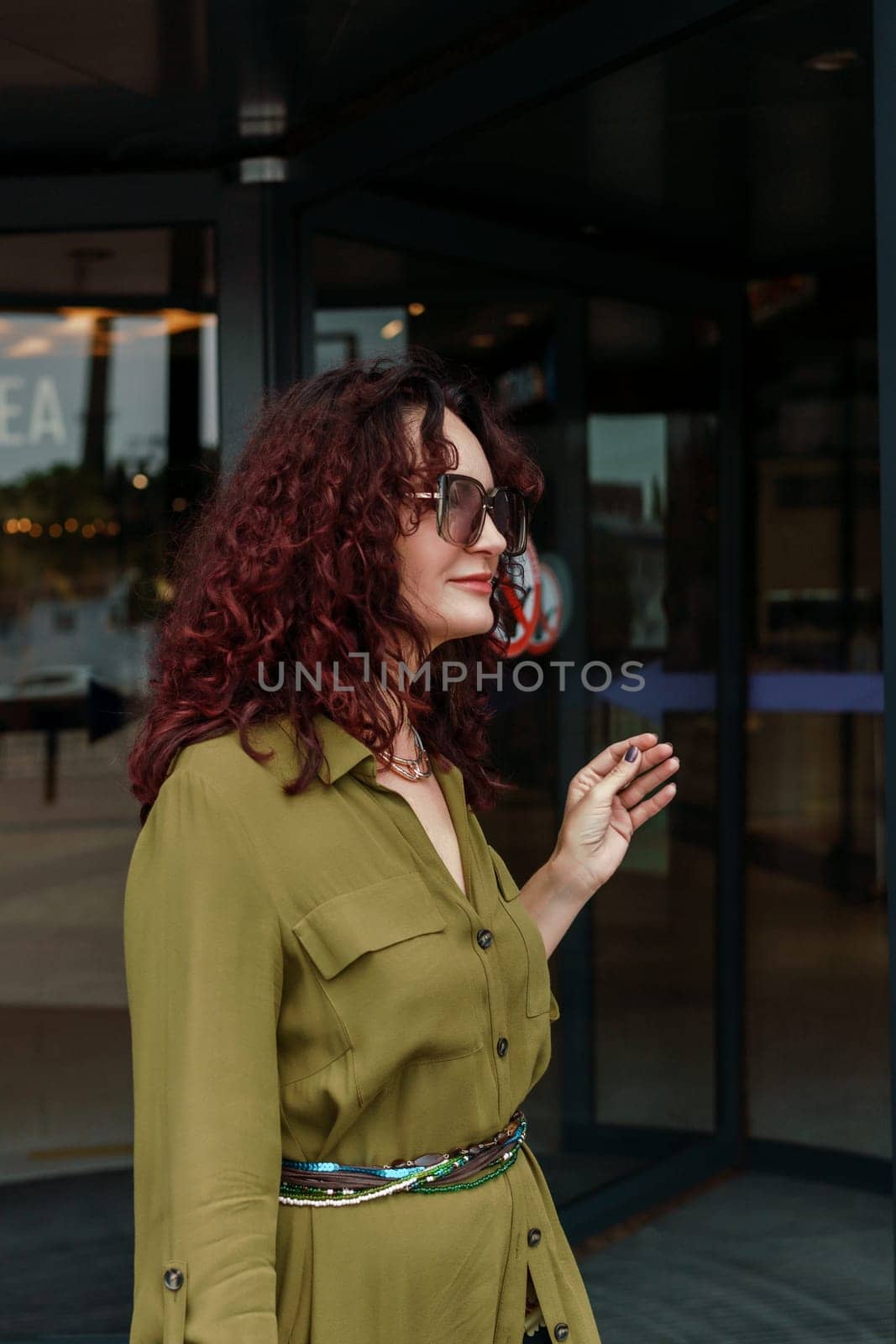 Woman leaves a supermarket. Caucasian model with long brunette hair, wears sunglasses and a khaki dress. by Matiunina