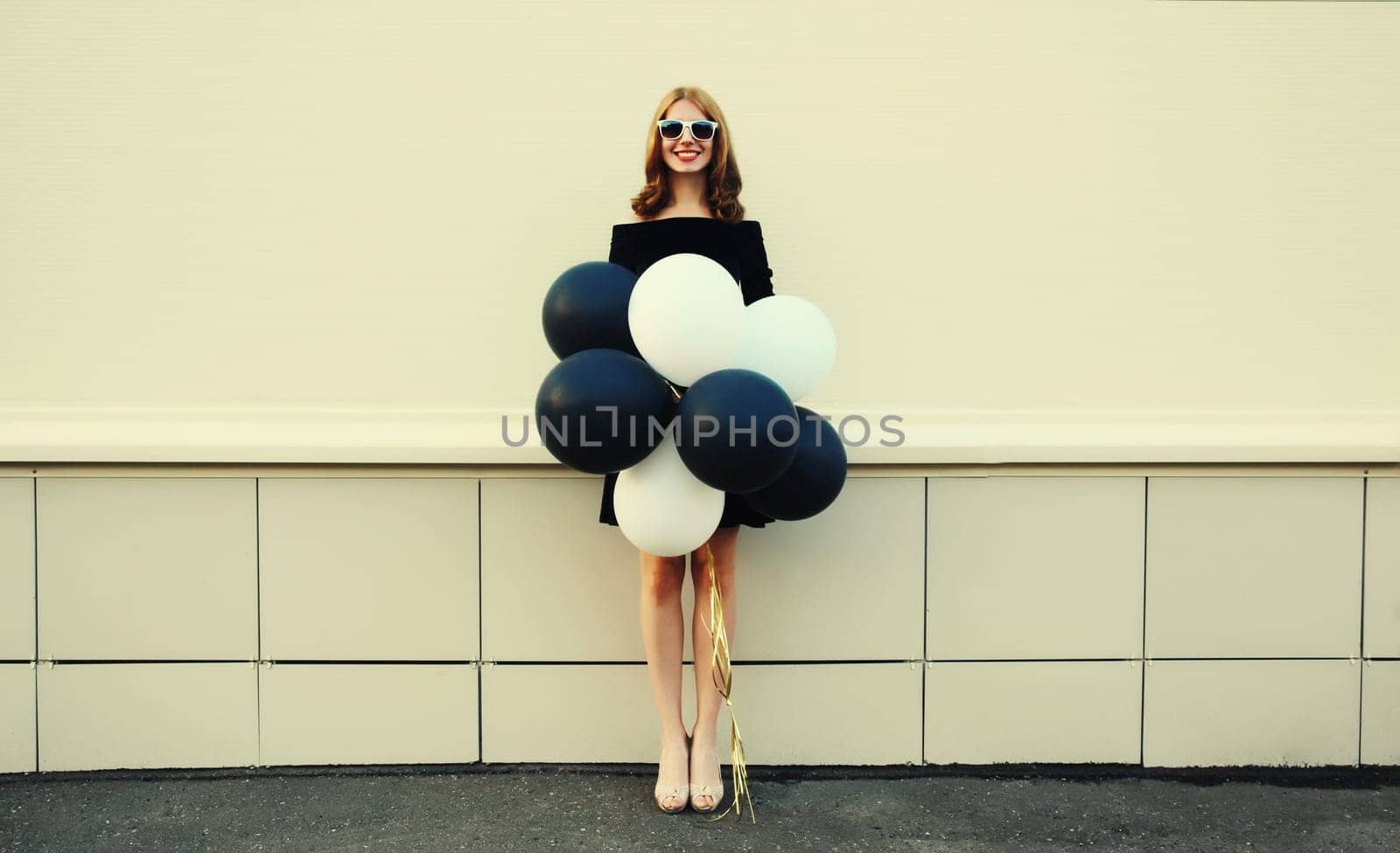 Beautiful happy smiling young woman full length with bunch of black and white balloons in summer dress on city street
