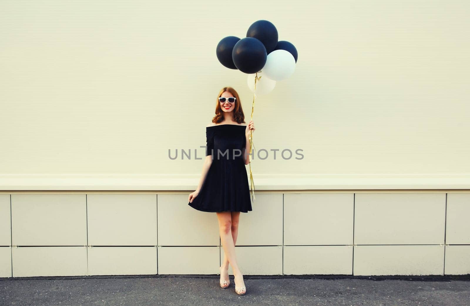 Beautiful happy smiling young woman full length with bunch of black and white balloons in summer dress on city street