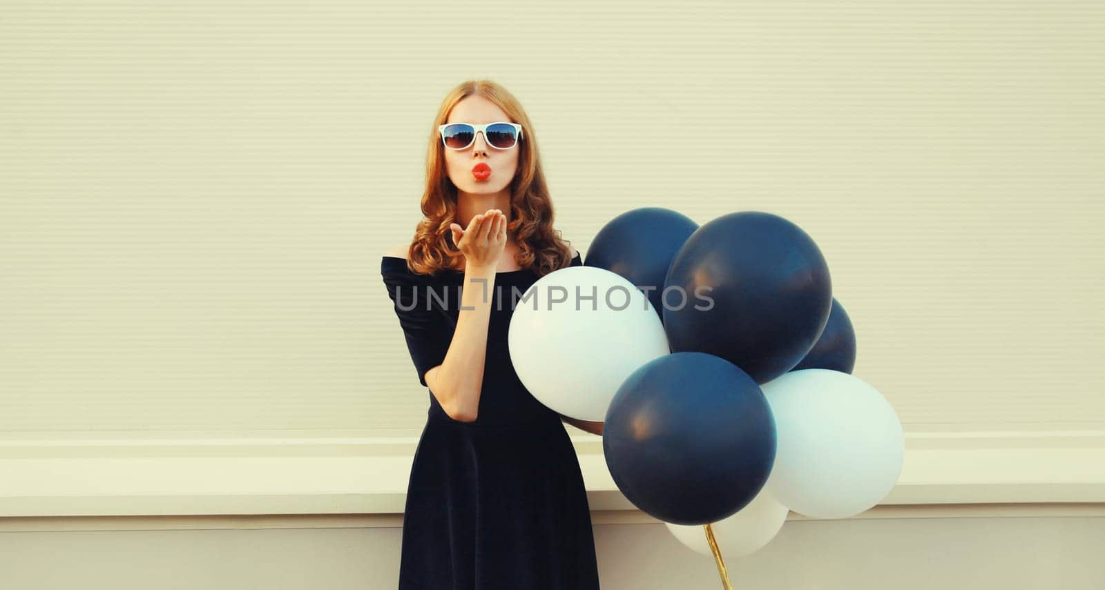 Portrait of beautiful happy young woman with bunch of black and white balloons in summer dress and blowing her lips sends kiss on city street
