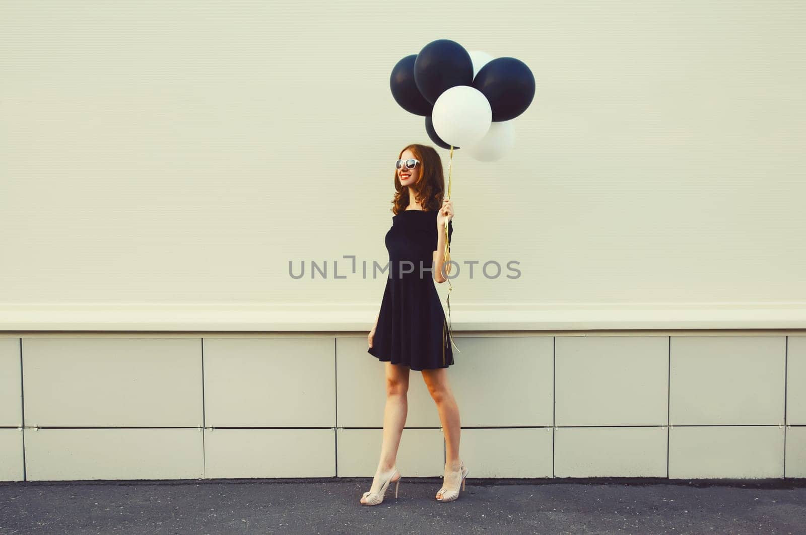 Beautiful happy young woman with bunch of black and white balloons in summer dress on city street by Rohappy