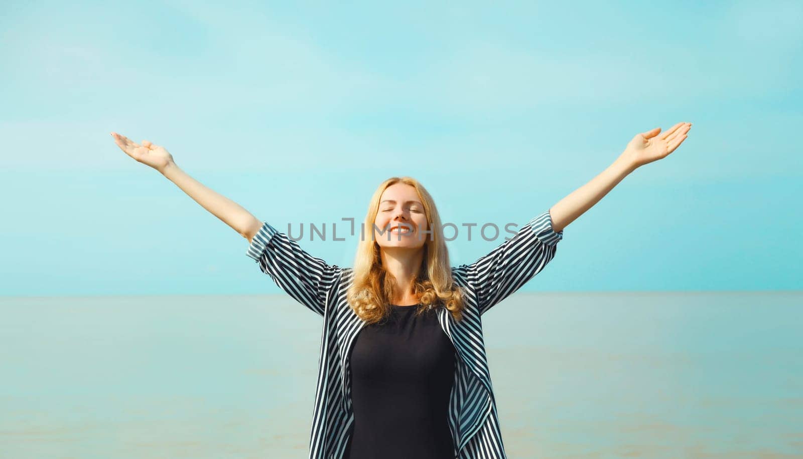 Summer vacation, happy young woman raising her hands up on the beach on sea coast and blue sky background