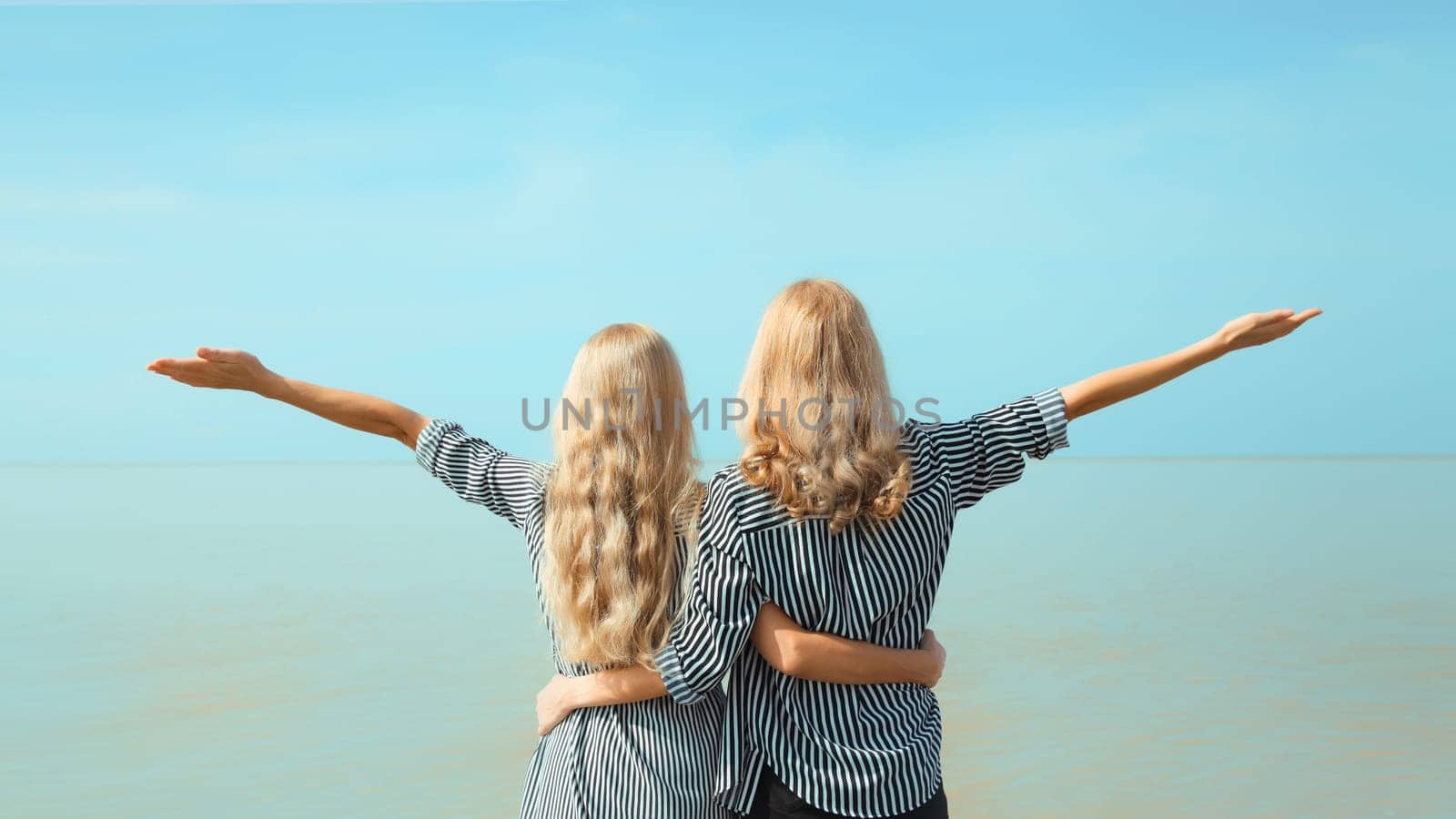 Summer vacation of two women raises her hands up on the beach, happy caucasian girlfriends or mother with adult daughter together on sea coast and blue sky background
