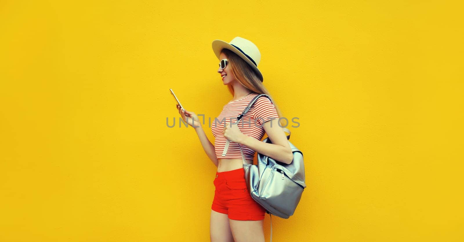 Summer portrait of happy traveler young woman 20s with mobile phone looking at device in casual straw hat with backpack on bright yellow studio background