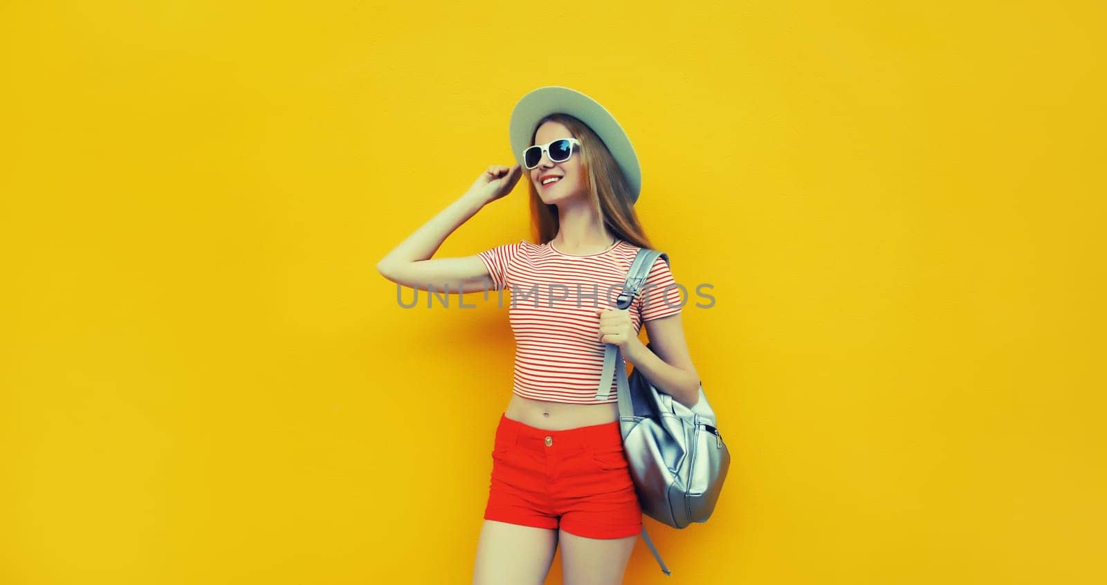 Beautiful young woman posing in summer straw hat, red shorts with backpack looking away on vivid yellow studio background