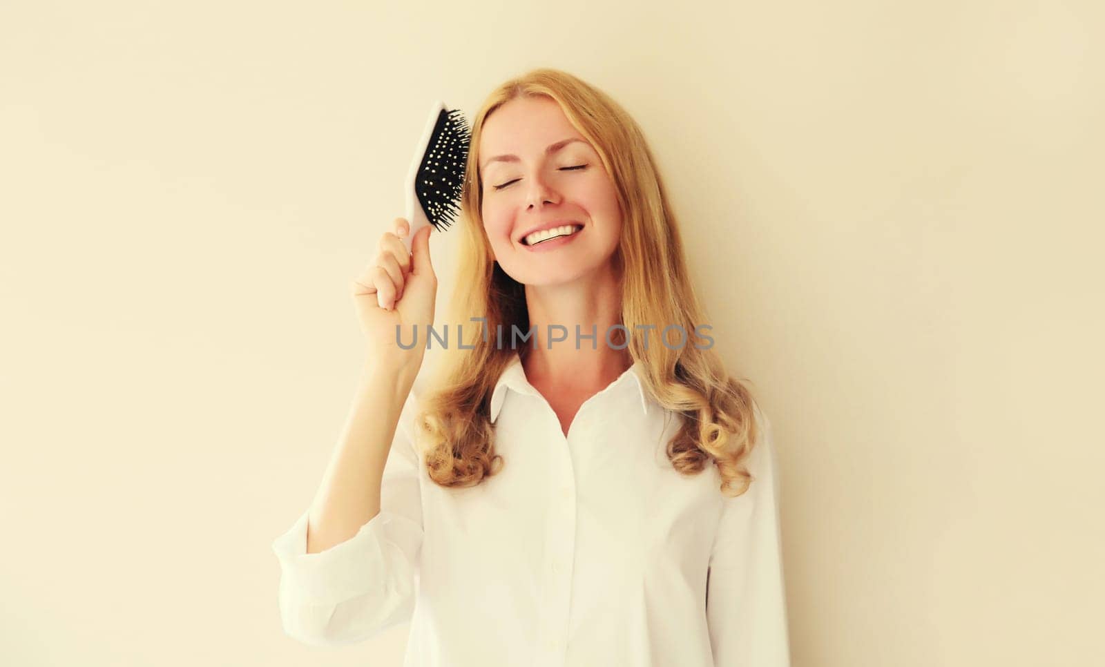 Portrait of happy smiling caucasian blonde young woman combing her hair with comb on white studio background