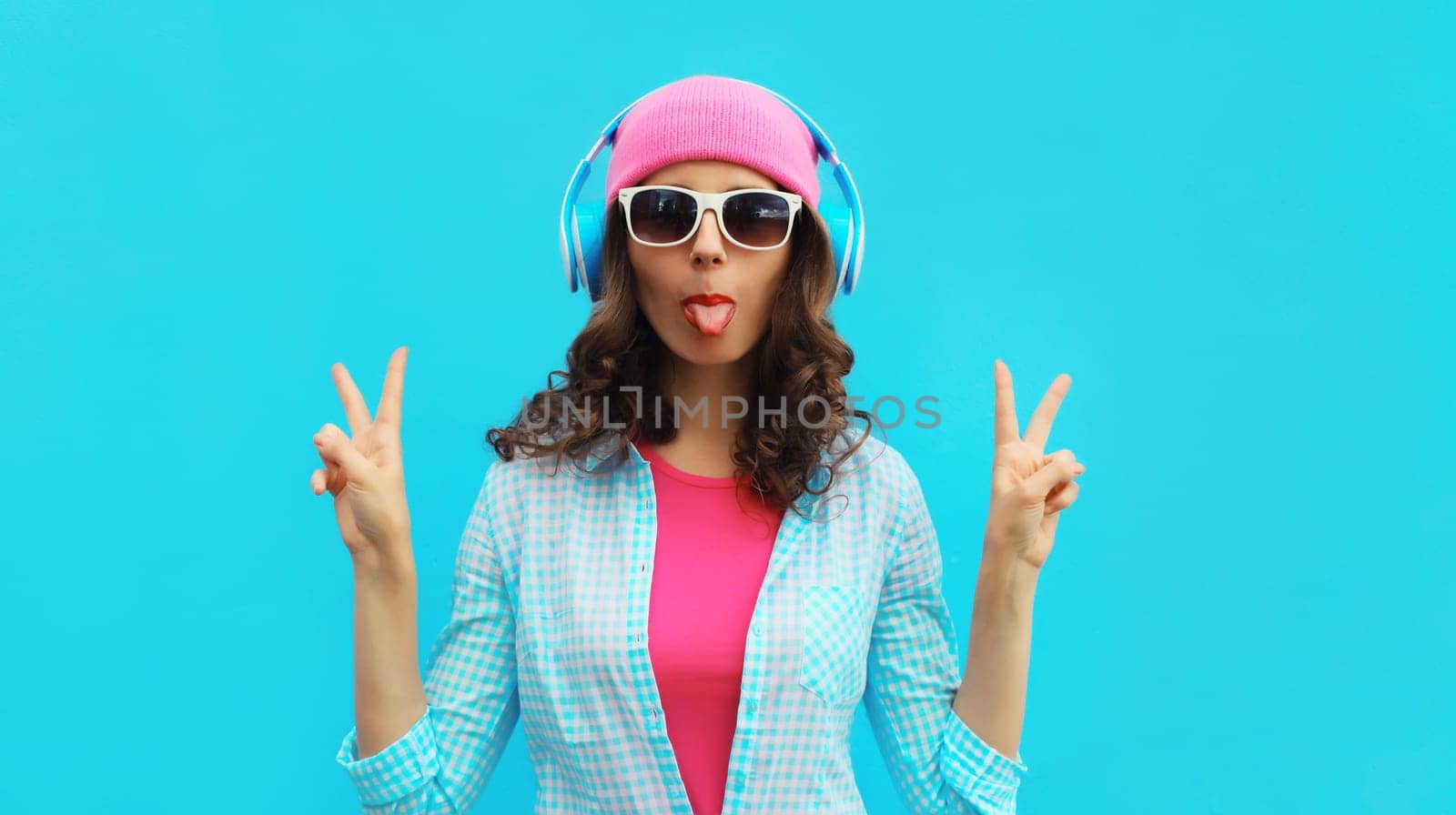 Portrait of stylish modern happy young woman listening to music with headphones wearing colorful pink hat on blue studio background