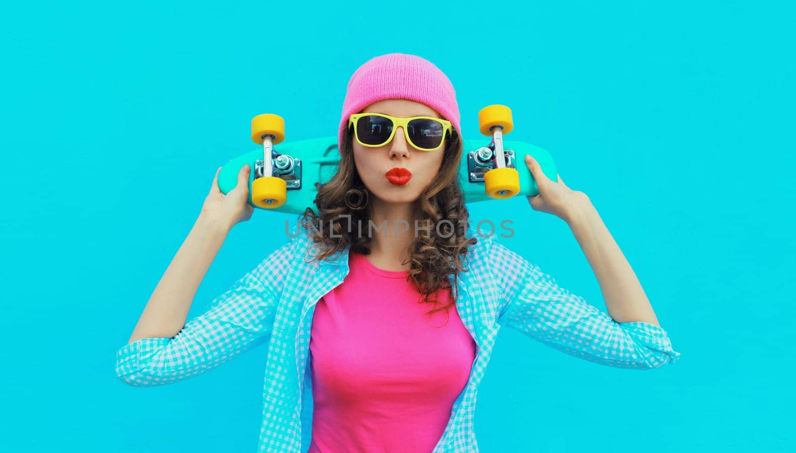 Portrait of stylish modern young woman with skateboard posing in colorful vivid clothes, pink hat on blue studio background