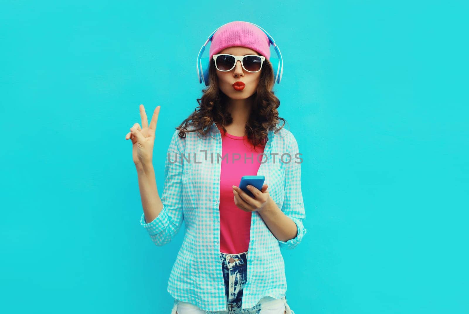 Portrait of stylish modern happy young woman listening to music in headphones with mobile phone wearing pink hat, sunglasses on blue studio background